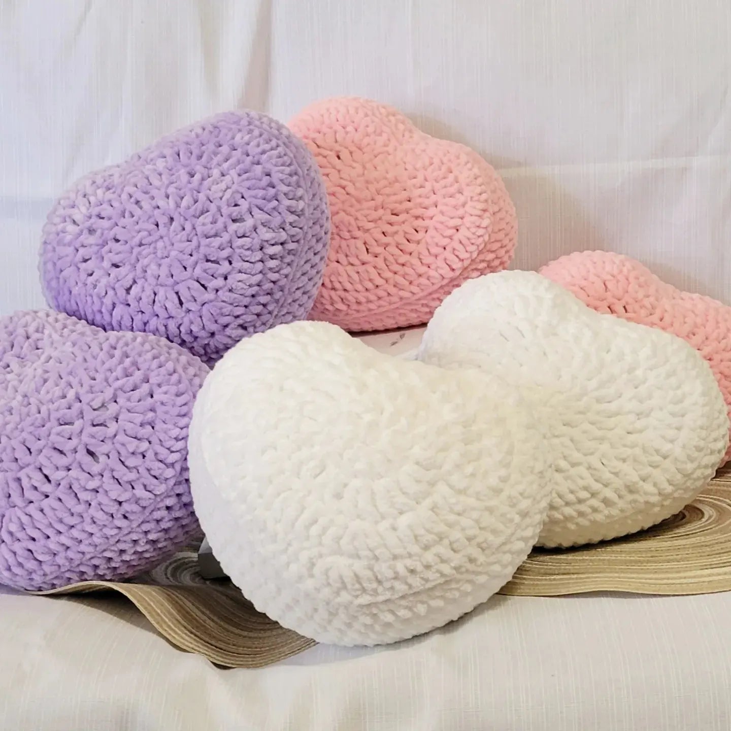 A pile crocheted heart pillows in purple, pink, and white on a white background