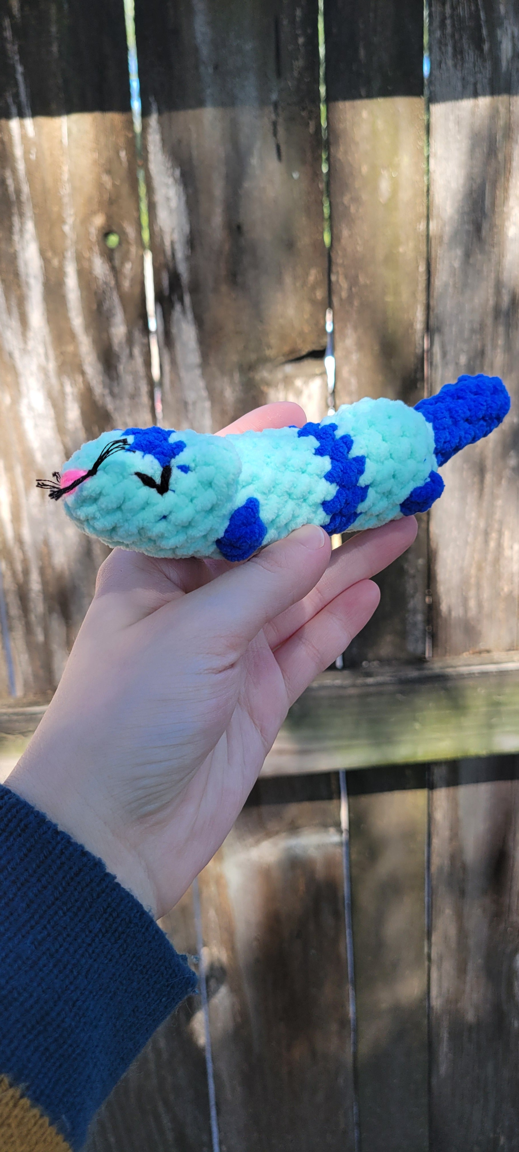Hand holding a blue and dark blue crocheted ferret toy against a wooden fence background