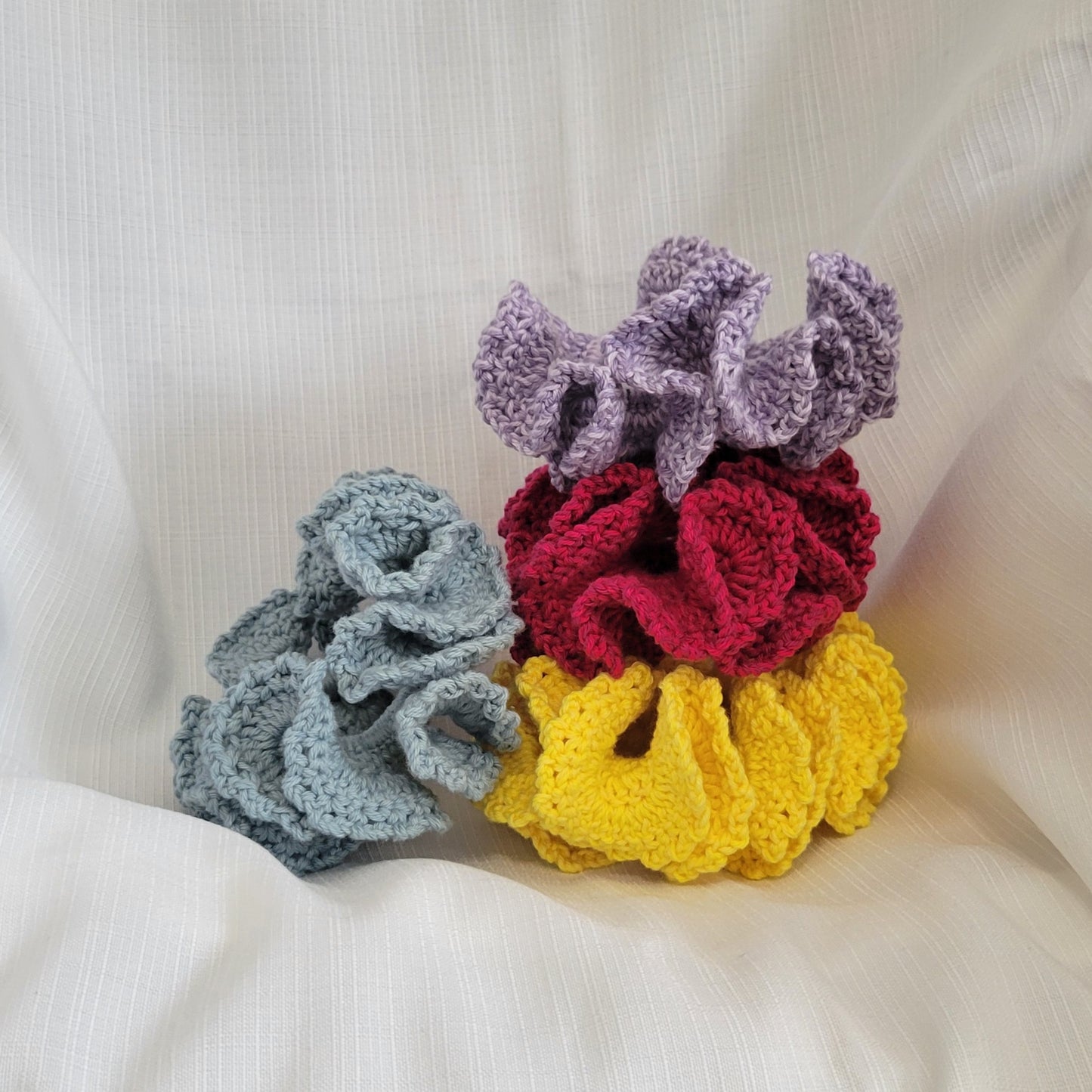 4 crocheted hair scrunchies stacked and 1 placed next to the stack. The colors are yellow, pinky red, purple and muted blue.