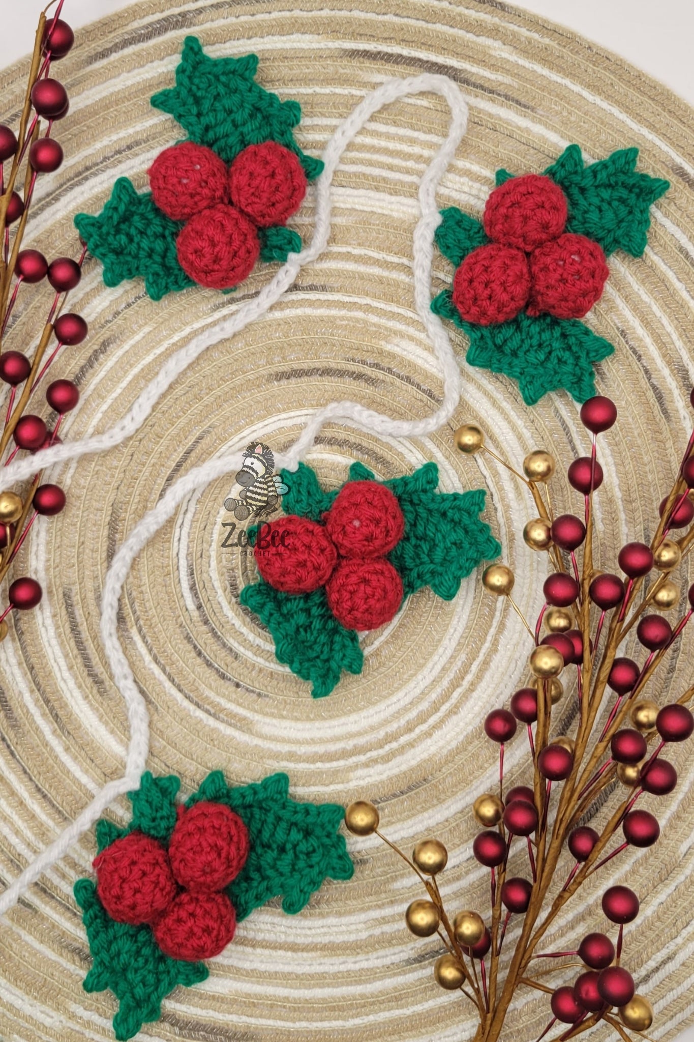 Strand of yarn chains that have holly berries and leaves attached on a wood grain background with decorative red and gold berries on stems.