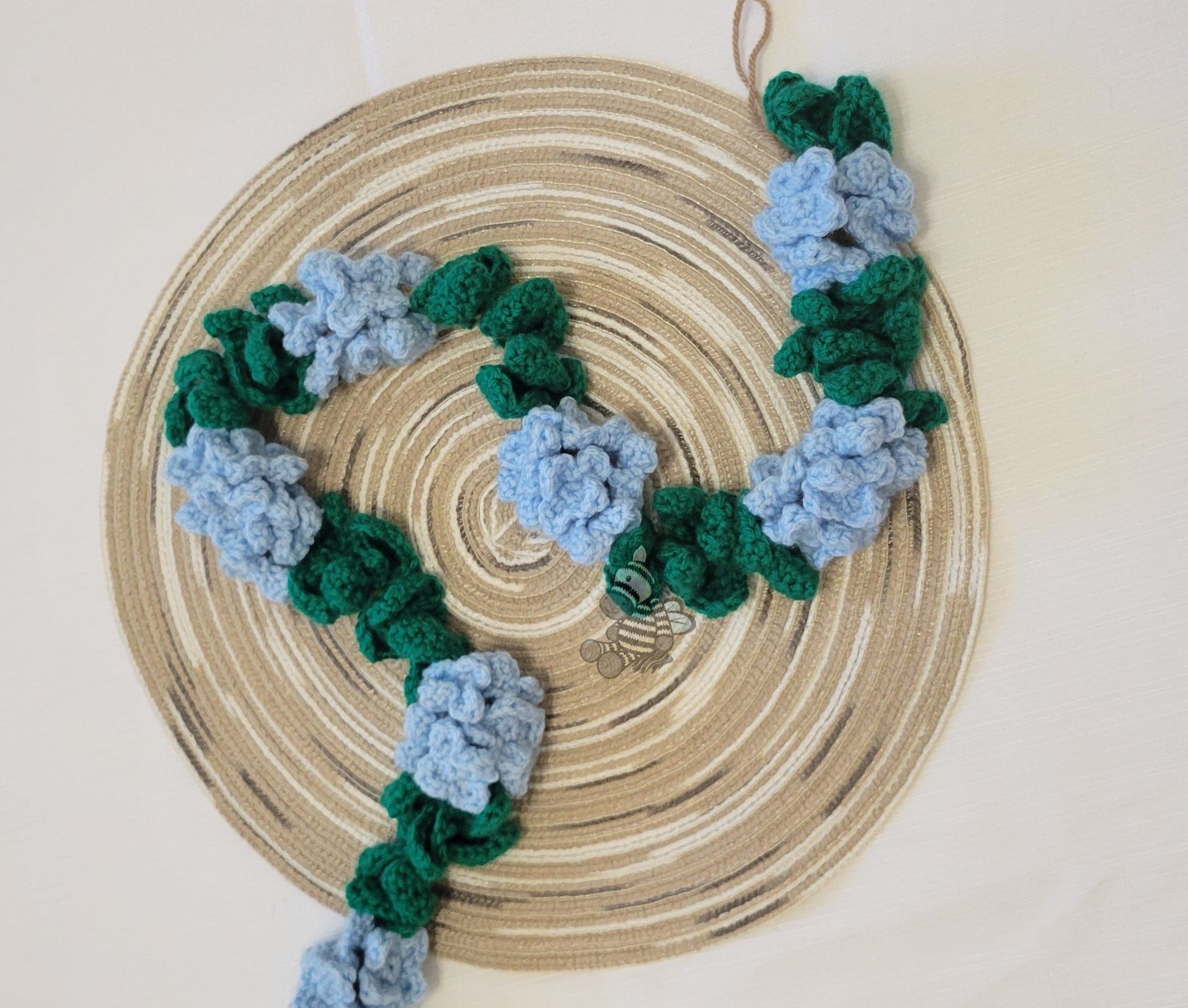 Garland with blue crochet hydrangea flowers and green leaves in a repeating pattern. The background is a wood grain circle and white backdrop.