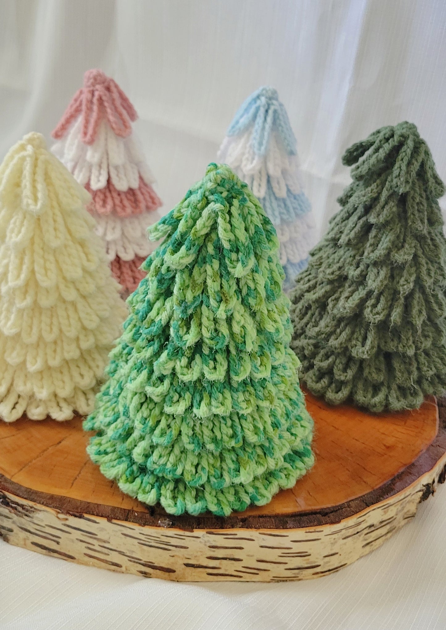 Crocheted Christmas trees in various colors on a wooden stand with a white background, colors are cream, white and pink and white and blue, dark green and multi green